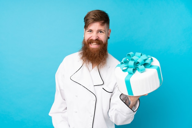 Chef de pastelaria ruiva com barba longa, segurando um bolo grande sobre parede azul isolada