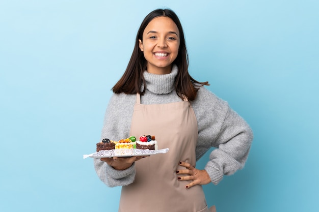 Chef de pastelaria segurando um bolo grande sobre fundo azul isolado, posando com os braços no quadril e sorrindo