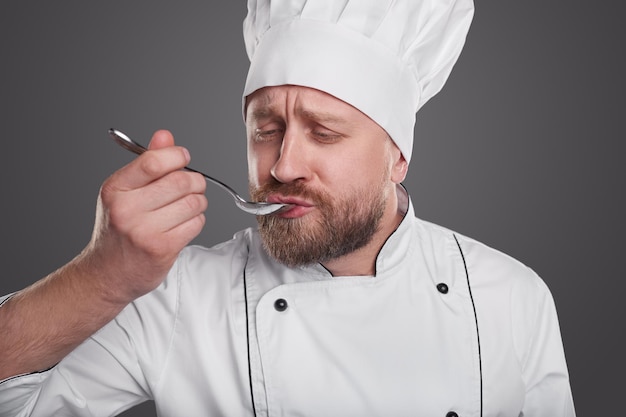Chef de uniforme branco degustando sopa quente com uma colher isolada sobre um fundo cinza