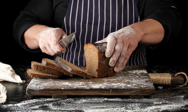 Chef de uniforme preto tem uma faca de cozinha na mão e corta pedaços de pão de um pão de farinha de centeio marrom assado