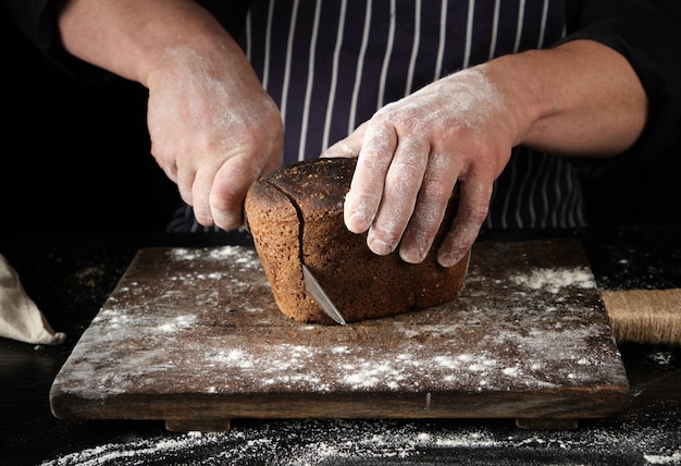 Chef de uniforme preto tem uma faca de cozinha na mão e corta pedaços de pão de um pão de farinha de centeio marrom assado