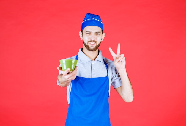 Chef en delantal azul sosteniendo una taza de cerámica verde y mostrando el signo de satisfacción.