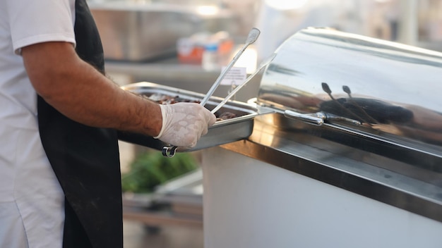 Chef em luvas de borracha segurando bandeja de metal com comida e pinças de perto