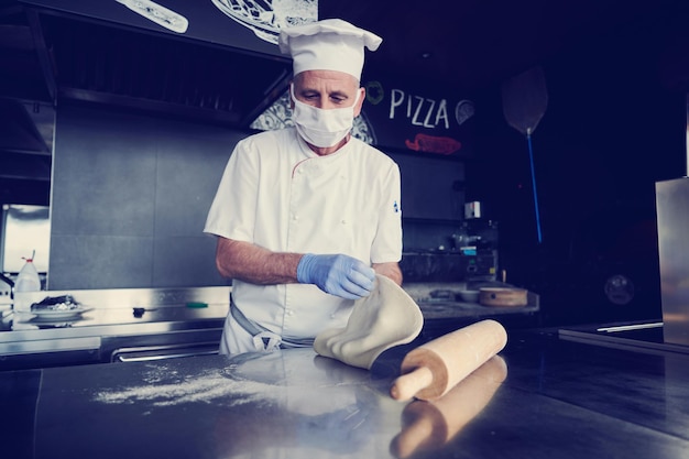 Chef experto que prepara pizza italiana tradicional en el interior de la cocina moderna del restaurante con horno de leña especial. Uso de mascarilla médica protectora y guantes en el nuevo concepto normal de coronavirus