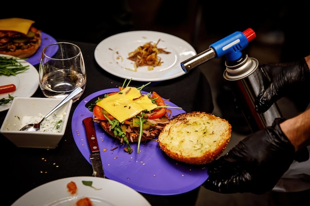 Chef fazendo hambúrgueres de carne derretendo queijo em um pão de hambúrguer com um queimador culinário para hambúrgueres em uma cozinha de restaurante