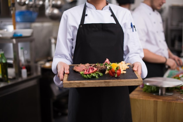 Chef femenina en la cocina del hotel o restaurante sosteniendo un plato de bistec de ternera a la parrilla con decoración vegetal