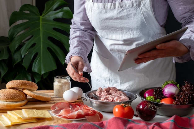 Foto chef feminino cozinhando big burger ou cheeseburger e usando tablet com tutorial de receita de culinária.