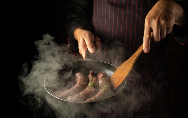 El chef fríe salchichas para perritos calientes en una sartén caliente con vapor