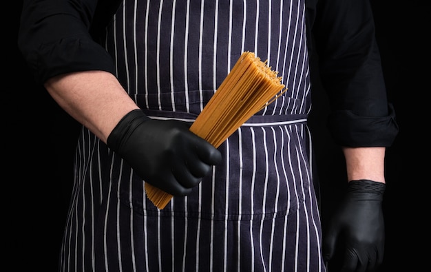 Foto chef con guantes de látex negro, delantal a rayas con espagueti amarillo crudo largo hecho de trigo duro