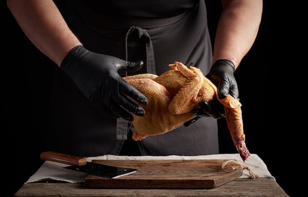 Chef en guantes de látex negro sostiene una carcasa de pollo entera sobre una tabla de cortar marrón, proceso de cocción de carne