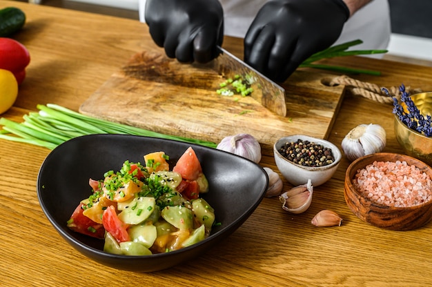 Un chef con guantes negros está cortando cebollas verdes frescas en una tabla de cortar de madera.