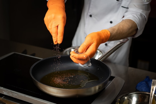 Un chef con guantes en sus manos agrega especias a la sartén con el plato de cocina