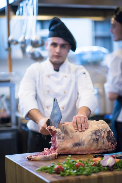 Chef con hacha mientras corta un gran trozo de carne de vacuno sobre una placa de madera en la cocina del restaurante