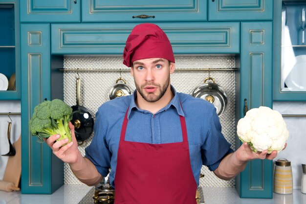 Chef hombre con sombrero rojo, delantal mantenga coliflor y brócoli vegetal en la cocina. Vegetariano, salud, concepto de dieta. Comida, cocina, culinaria, gastronomía.