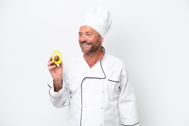 Chef hombre sosteniendo un aguacate aislado sobre fondo blanco mirando hacia un lado y sonriendo