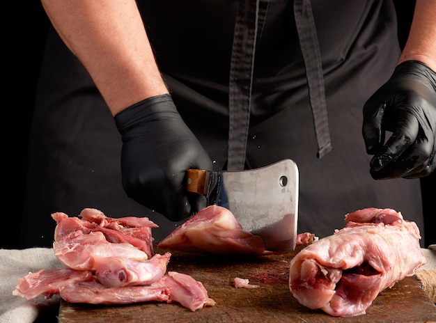 Chef in schwarzen Latexhandschuhen hält ein großes Messer und schneidet rohes Kaninchenfleisch in Stücke