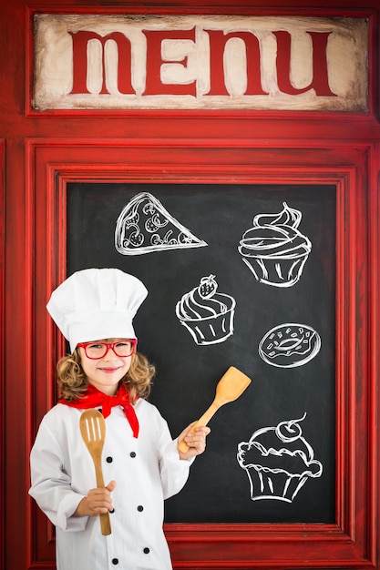 El chef infantil cocina contra el menú en blanco de la pizarra con el dibujo de alimentos saludables. Concepto de negocio de restaurante