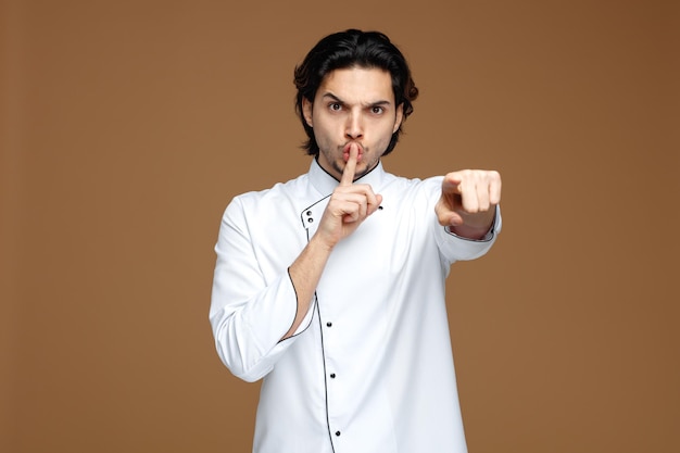 Un chef joven y estricto con uniforme mirando y apuntando a la cámara mostrando un gesto de silencio aislado de fondo marrón