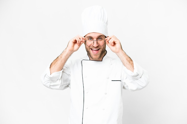 Chef joven rubio sobre fondo blanco aislado con gafas y sorprendido