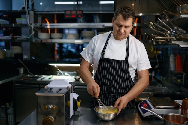 Chef maduro con delantal batiendo huevo en un tazón mientras cocina pasta en la cocina del restaurante. receta culinaria para el desayuno