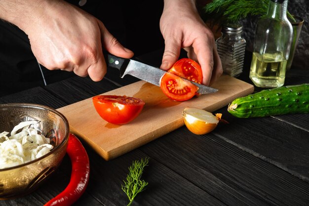 Chef mãos com faca, cortando tomates vermelhos frescos para salada. Ambiente de trabalho na cozinha do restaurante. Legumes frescos e óleo na mesa