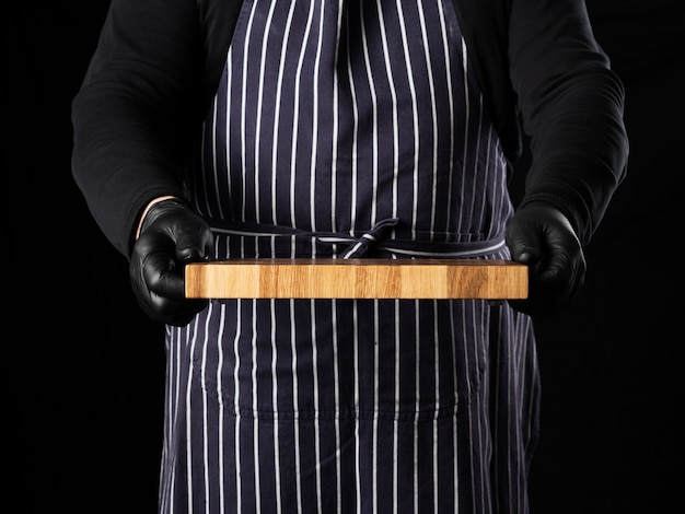 Chef masculino com um avental listrado azul e roupas pretas fica contra um fundo preto e segura na mão uma tábua de cozinha retangular de madeira, close-up