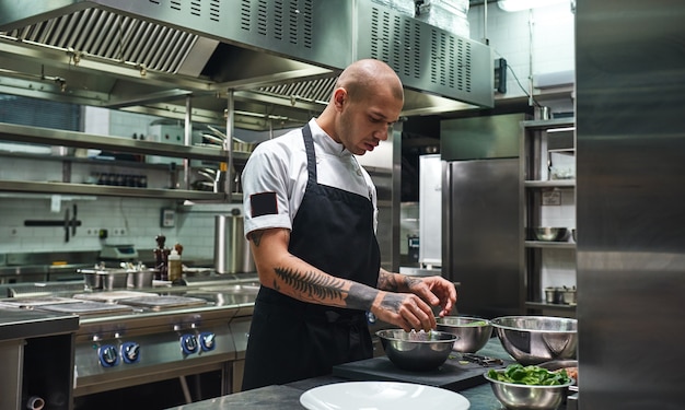 Chef masculino de avental, com tatuagens pretas nos braços, preparando salada na cozinha de um restaurante