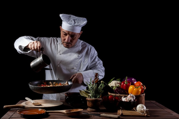 Chef masculino de uniforme branco e chapéu servindo azeite de oliva na frigideira com legumes antes de servir enquanto trabalha na cozinha de um restaurante