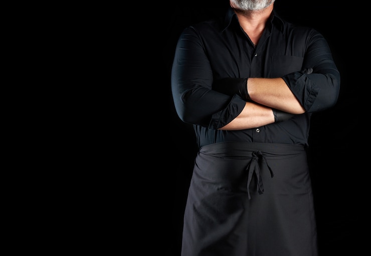 Foto chef masculino de uniforme preto cruzou os braços na frente do peito em um fundo preto, banner para restaurantes e cafés, espaço vazio para uma inscrição