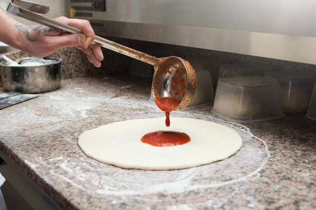 Chef masculino derrama molho vermelho na pizza pré-forma de massa crua produção e entrega de pizza