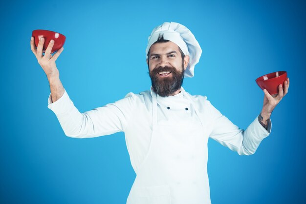 Chef masculino em uniforme branco segura tigelas vermelhas chef barbudo isolado no conceito de cozinha de fundo azul