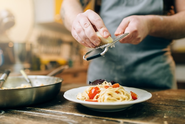 Chef masculino ralla queso en el plato con pasta fresca cocida, pan en la mesa de la cocina de madera. Proceso de preparación de fettuccine casero. Cocina tradicional italiana