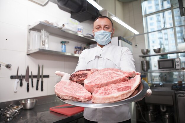 Chef masculino usando máscara médica e segurando um prato com carne crua em mármore