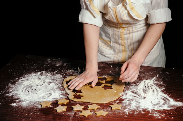 Chef mujer en delantal hace galletas en forma de estrellas de masa sobre mesa de madera con harina