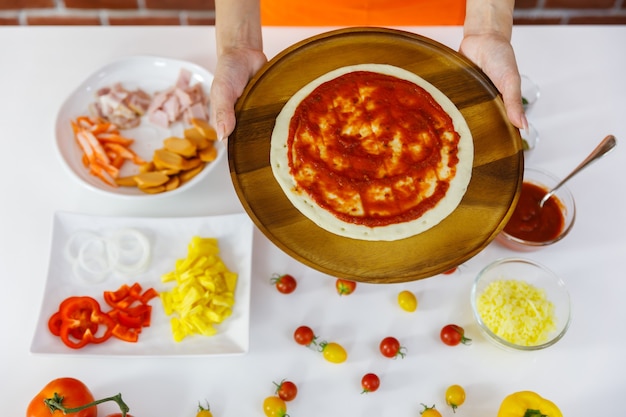 Chef mujer en delantal naranja preparándose para cocinar sabrosa pizza saludable en plato de madera con variedad de tomates frescos rojos y amarillos, carne, ingredientes orgánicos y condimentos en la mesa blanca cerca de la pared de ladrillo