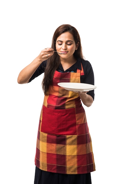 Chef mujer india vistiendo delantal y sosteniendo un plato y un tazón de cerámica vacíos con diferentes expresiones faciales que se encuentran aisladas sobre fondo blanco.