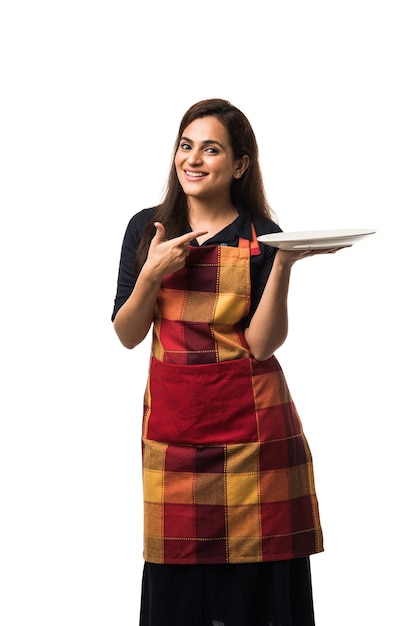 Chef mujer india vistiendo delantal y sosteniendo un plato y un tazón de cerámica vacíos con diferentes expresiones faciales que se encuentran aisladas sobre fondo blanco.