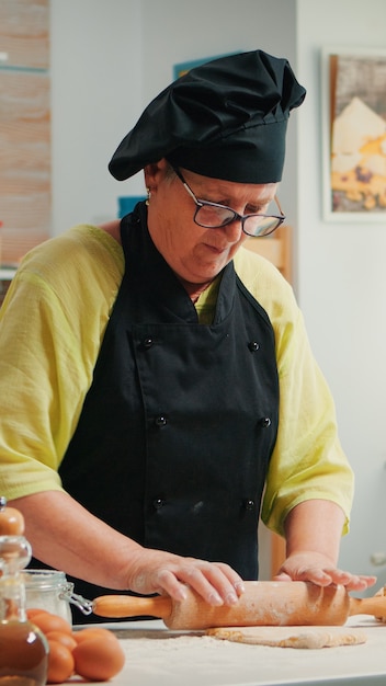 Chef mujer jubilada con rodillo de madera para cocinar pizza. Feliz panadero anciano con bonete preparando ingredientes crudos para hornear pizza tradicional espolvoreando, tamizando la harina en la mesa de la cocina.