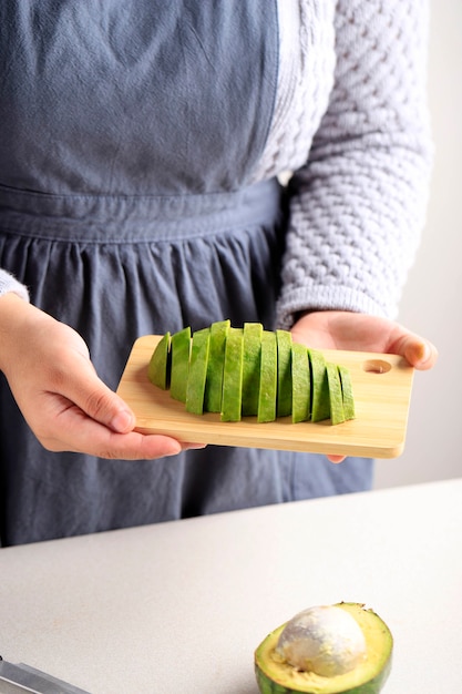 Chef mujer mantenga rodajas de aguacate sobre tabla de madera, cocinando entre bastidores en la cocina haciendo comida sana