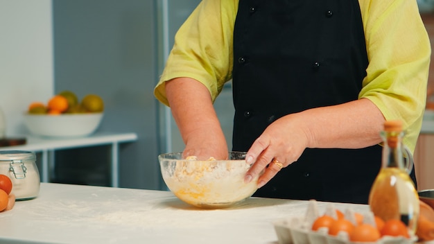 Chef mujer mayor mezclando a mano los ingredientes del pan en la cocina casera en un tazón de vidrio. Anciano panadero jubilado con bonete amasando huevos frescos agrietados con harina de trigo para hornear pan y pasteles caseros