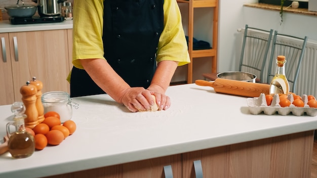 Chef mujer senior amasa la masa sobre la mesa en la cocina moderna. Anciano panadero jubilado con bonete mezclando ingredientes con harina de trigo tamizada amasando para hornear pasteles y pan tradicionales.