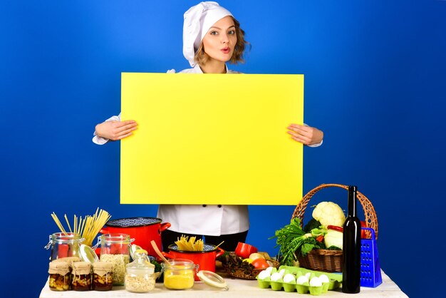 Chef mulher detém placa amarela para publicidade de texto. Preparo da comida. Chef sorridente, sente-se à mesa. Copie o espaço para publicidade de restaurante. Menu saudável. Dieta. Estilo de vida saudável.