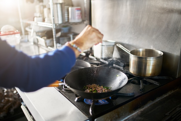 Chef na cozinha do restaurante temperando comida em frigideira quente wok