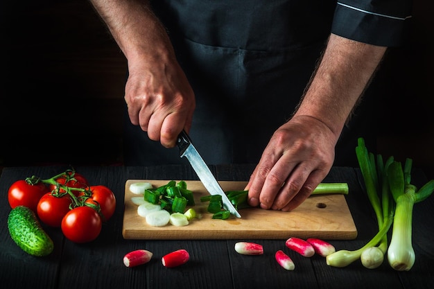 Chef oder Koch ist Hände Nahaufnahme schneidet junge Frühlingszwiebeln auf einem Schneidebrett der Restaurantküche für Salat. Gemüsediät oder Snackidee.