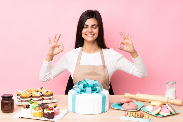 Chef pastelero con un gran pastel en una mesa sobre pared rosa aislado en pose zen