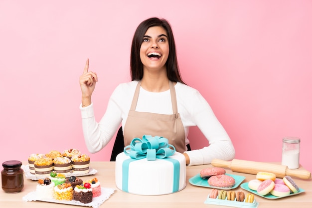 Chef pastelero con un gran pastel en una mesa sobre pared rosa apuntando hacia arriba y sorprendido