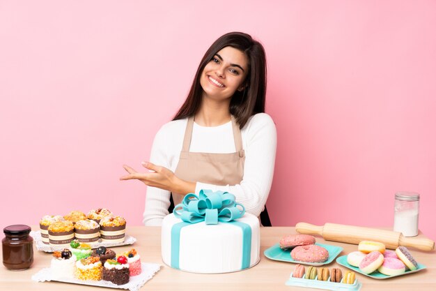 Chef pastelero con un gran pastel en una mesa sobre la pared rosa presentando una idea mientras mira sonriente hacia