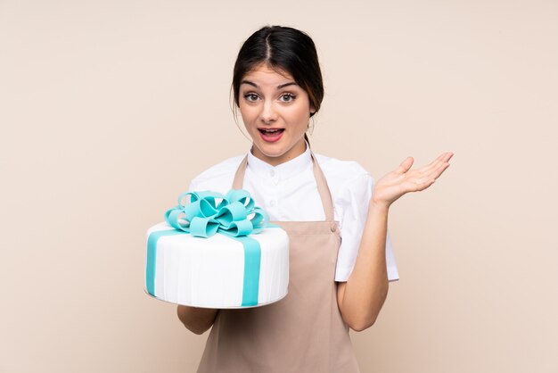 Chef pastelero mujer sosteniendo un gran pastel sobre la pared con expresión facial sorprendida