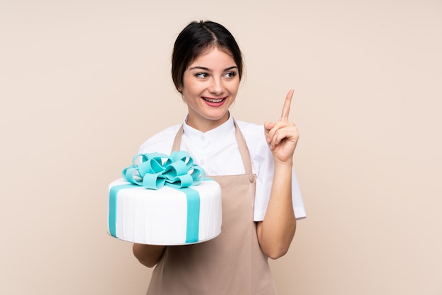 Chef pastelero mujer sosteniendo un gran pastel sobre la pared con la intención de darse cuenta de la solución mientras levanta un dedo