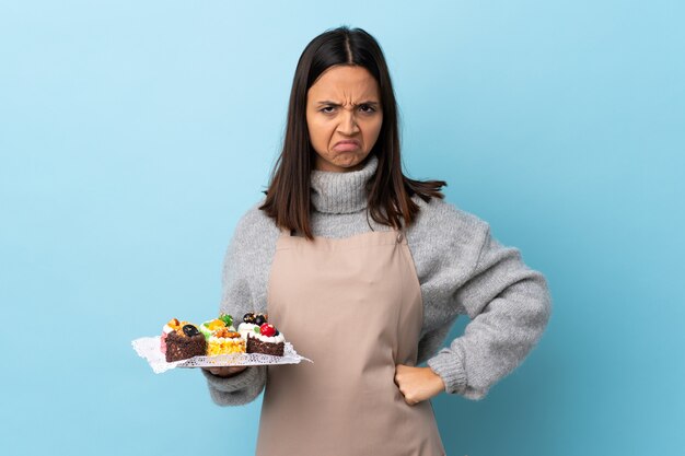 Chef pastelero sosteniendo un gran pastel sobre azul aislado con expresión infeliz.